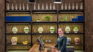 Eine Frau steht in einem Raum mit Holzboden, einem Schreibtisch mit Computer und Regalen mit diversen Gegenständen.