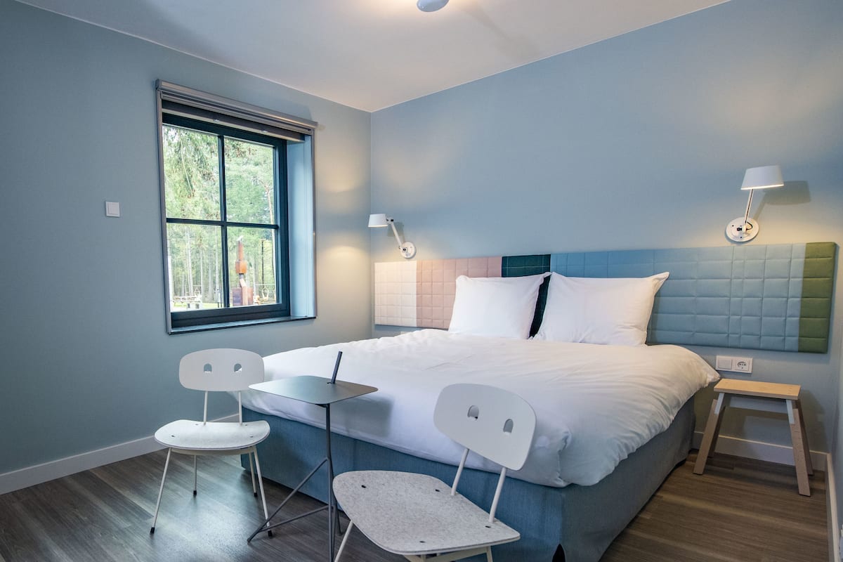 A modern bedroom with a bed, two chairs, a small table, and a window.