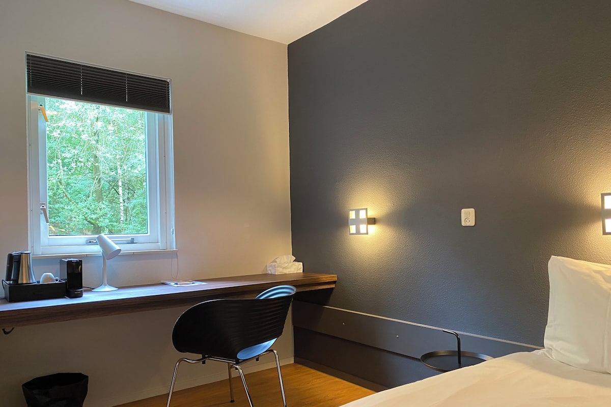 A bedroom with a bed, a desk with a chair, a window with blinds, and two wall-mounted lights.