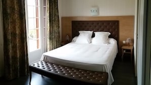 Ein ordentlich bezogenes Bett mit weißer Bettwäsche, ein Holzkopfstück und eine Sitzbank am Fußende. Ein Nachttisch mit Lampe und eine Wanduhr sind ebenfalls vorhanden.