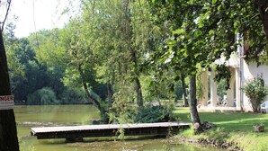 Eine friedliche Flusslandschaft mit einer hölzernen Anlegestelle, die sich in ruhiges Gewässer erstreckt, umgeben von üppigem Grün und einem Gebäude mit Säulen im Hintergrund.