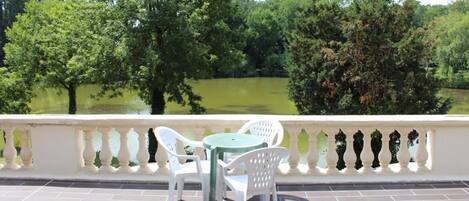 A terrace with white chairs and a table, overlooking a pond surrounded by trees.