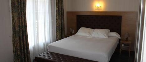 Quarto de hotel com cama grande, um banco, mesa de cabeceira com abajur e luminária na parede.