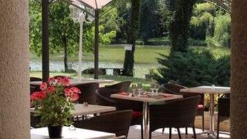 Un patio al aire libre con mesas y sillas, un área de estar cubierta y vistas a un lago y follaje.