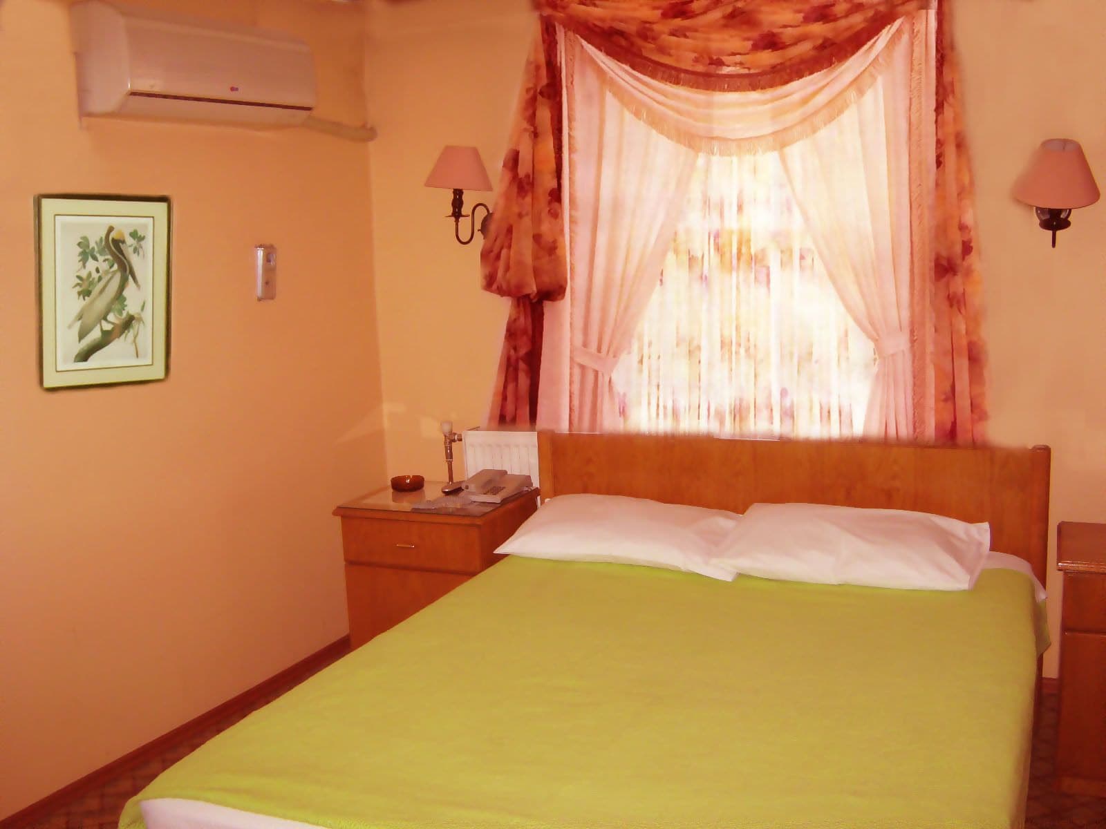 A hotel room with a bed, a nightstand, an air conditioner, a framed picture, and two wall lamps.
