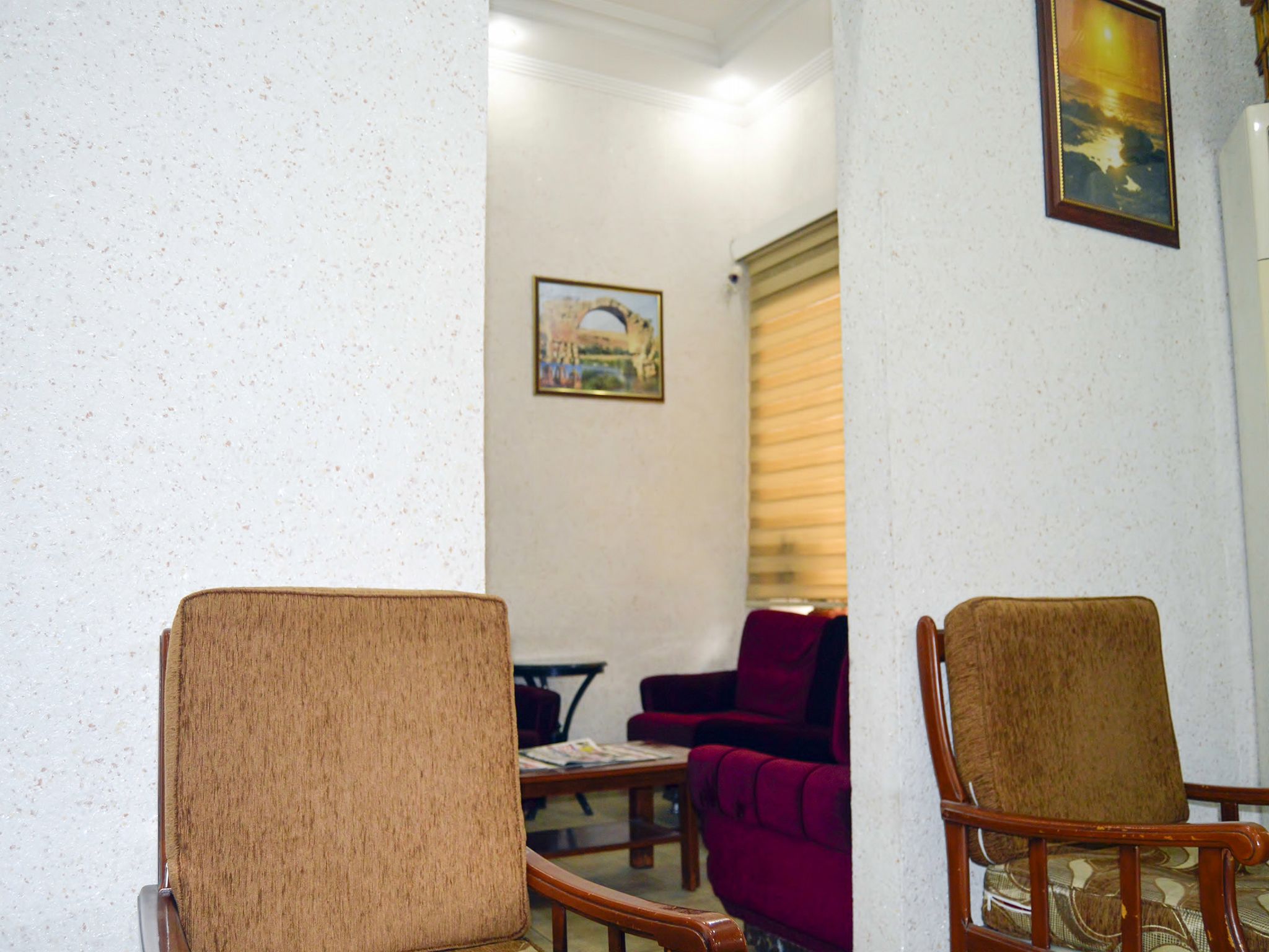 Uma sala de estar com duas cadeiras de madeira, um sofá vermelho e uma mesa. Há um quadro na parede e uma janela com persianas.