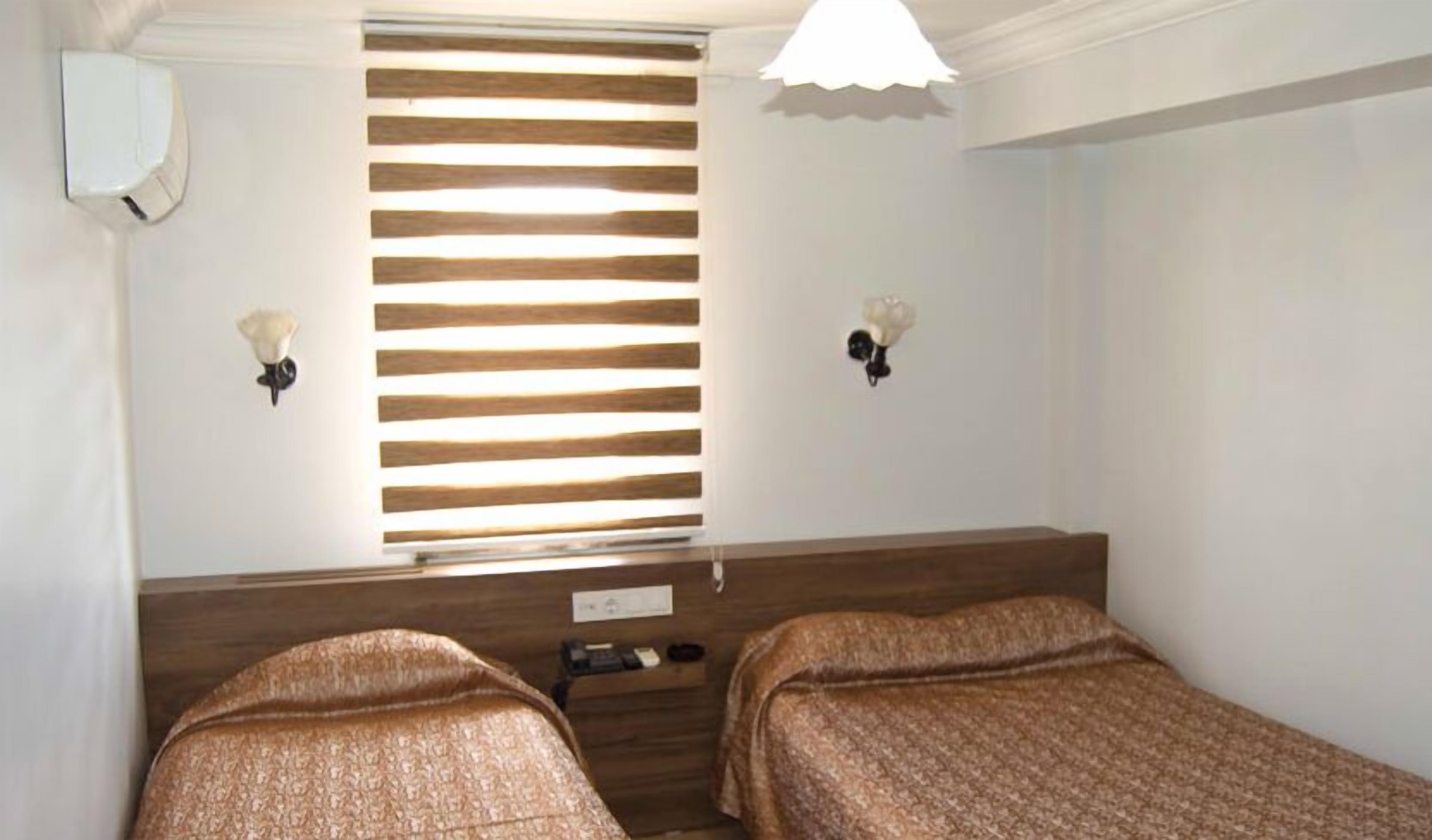 A room with two beds, a window with horizontal blinds, a white ceiling light, and wall-mounted lamps.