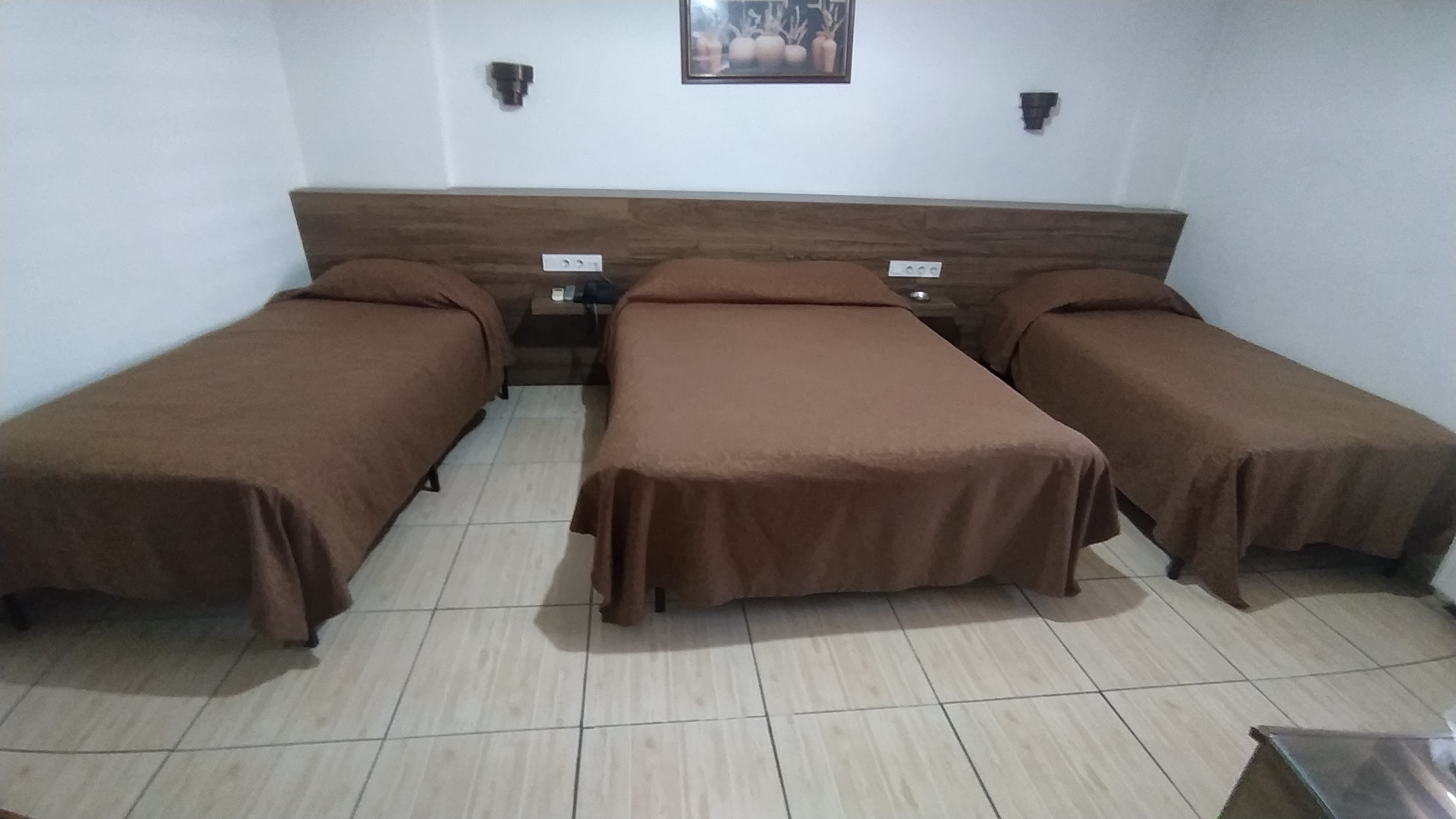 A room with three single beds, each with a brown bedspread, a wooden headboard, and a small bedside table with a lamp. There are two wall-mounted light fixtures and a framed picture on the wall.