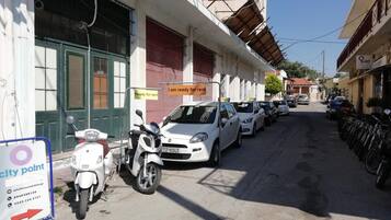 Una calle estrecha bordeada de coches y scooters aparcados, un cartel que indica disponibilidad de alquiler, y un edificio con una persiana roja y un balcón.