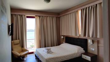 A hotel room with a bed, a chair, a bedside table, and a view of the sea through large windows.