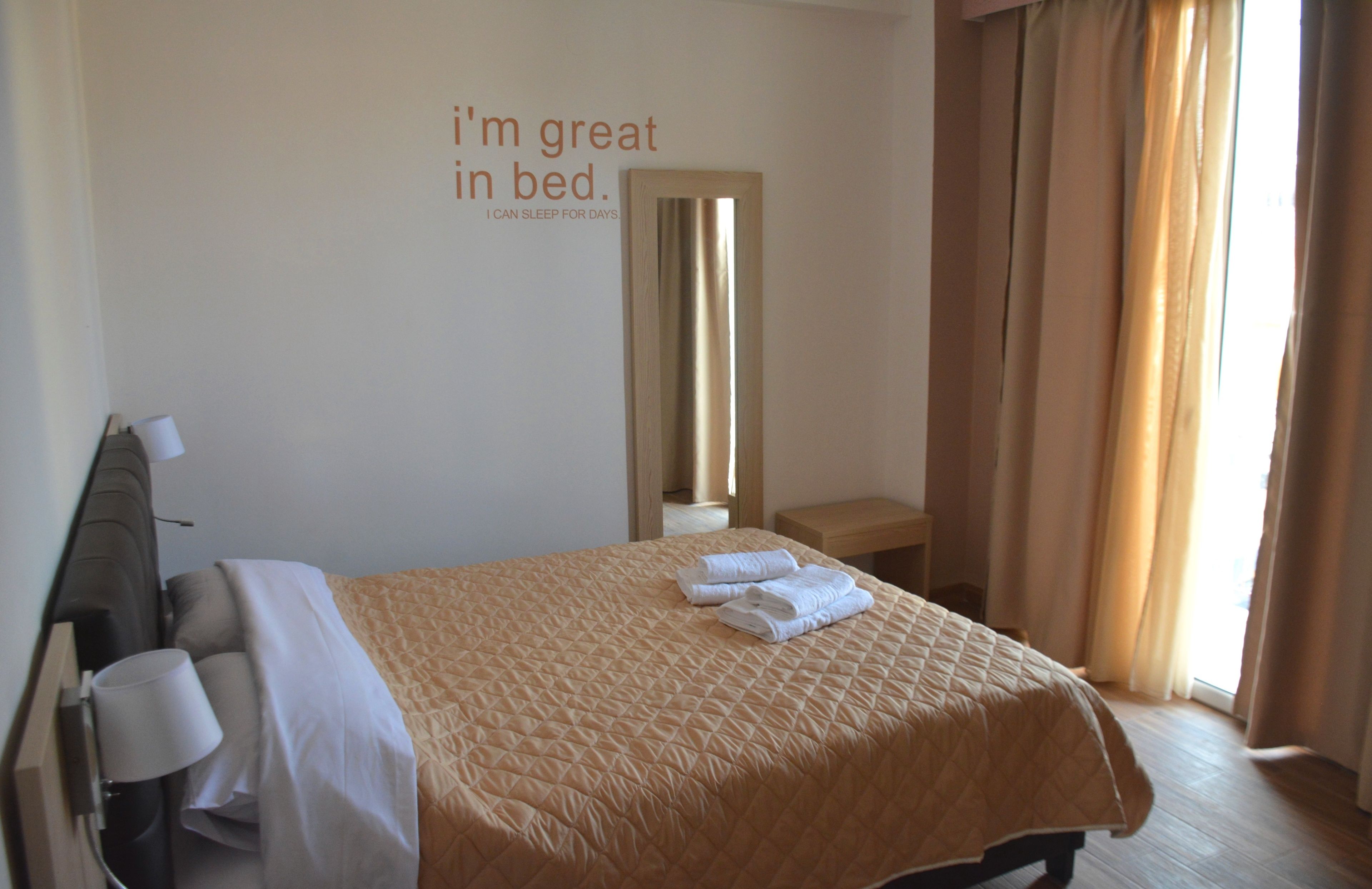 A hotel room with a bed, a bedside table, a mirror, and a wall with a motivational quote.