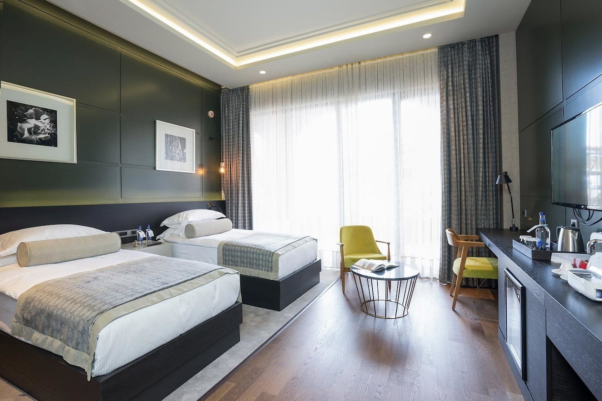 A hotel room with two beds, a desk with a chair, a TV, and a large window with curtains.