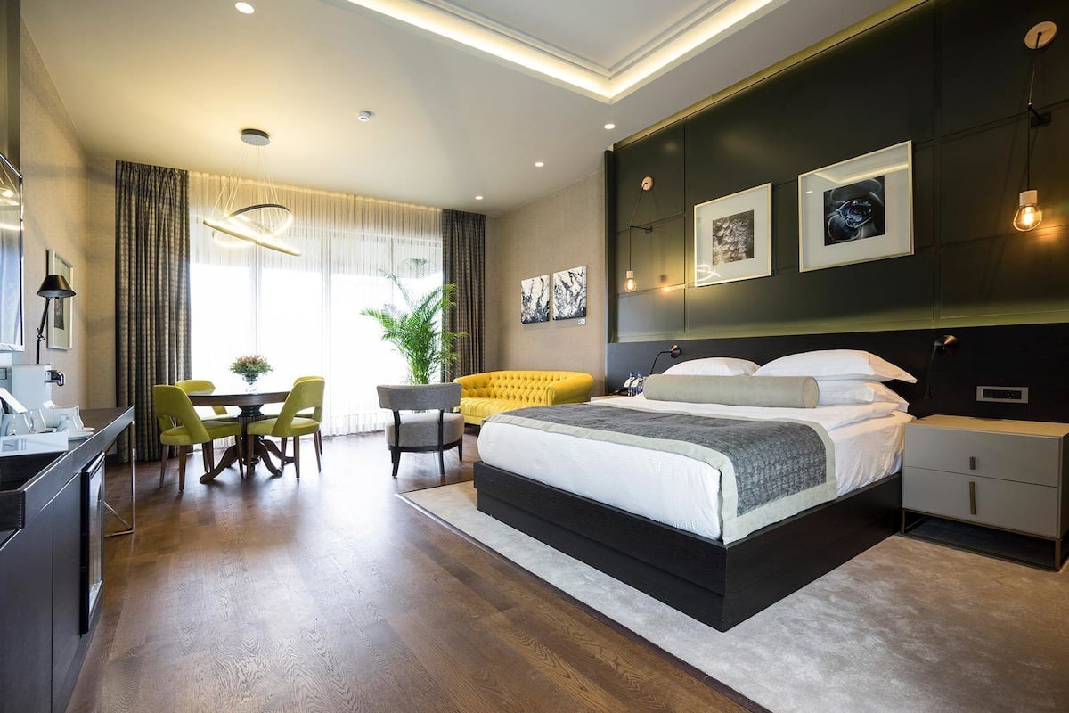 A modern hotel room with a large bed, a dining area with yellow chairs, and a desk with a lamp.