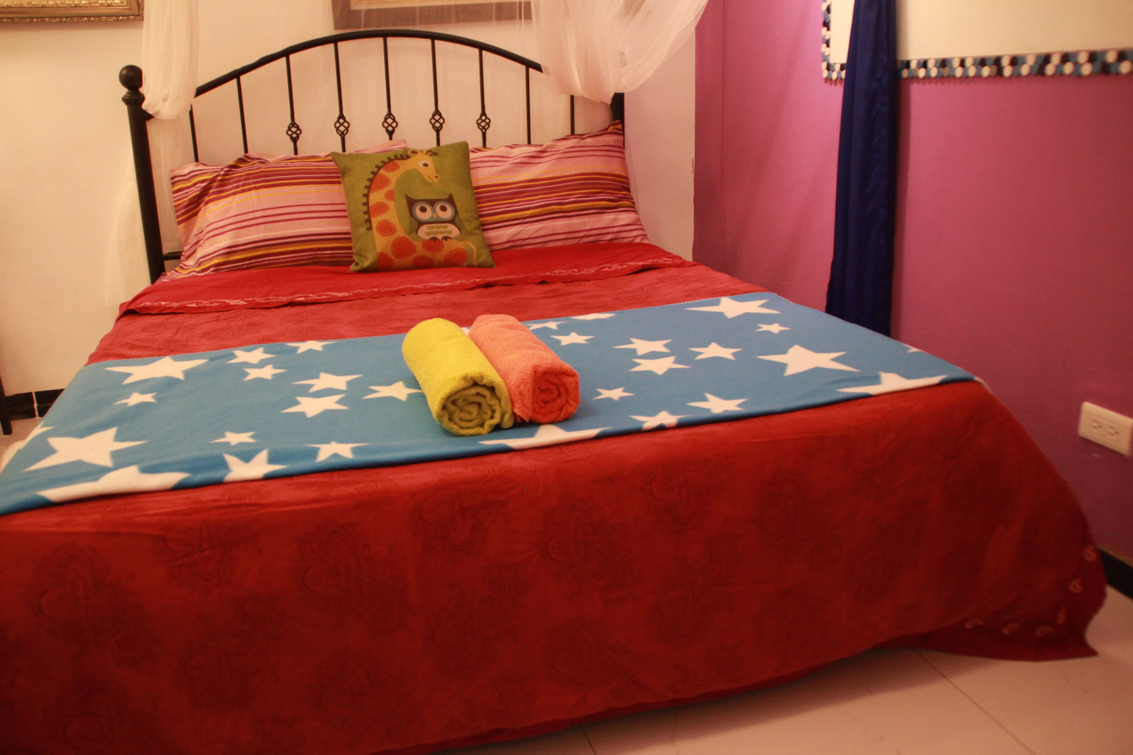 Una cama con una colcha roja y azul adornada con estrellas blancas, un almohadón verde con diseño de lechuza y dos toallas enrolladas en tonos amarillo y naranja.