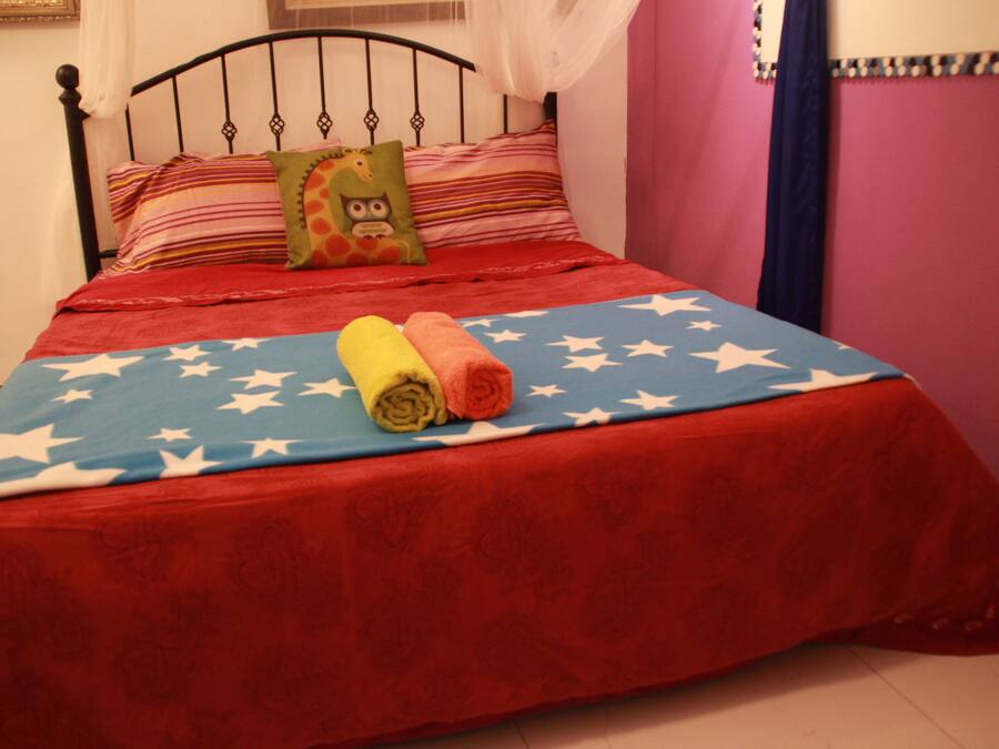 Una cama con una colcha roja y azul adornada con estrellas blancas, un almohadón verde con diseño de lechuza y dos toallas enrolladas en tonos amarillo y naranja.