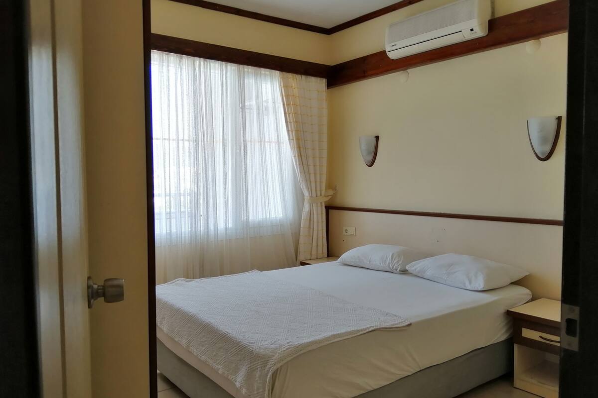 A bedroom with a bed, a nightstand, a wall-mounted air conditioner, and a window with sheer curtains.