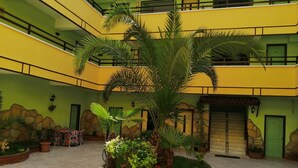 A courtyard with a central palm tree surrounded by greenery, a brick wall, and a building with yellow and green walls. - Oya Apartments (Kusadasi)