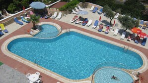 A large rectangular swimming pool with a circular section, surrounded by lounge chairs and umbrellas. - Oya Apartments (Kusadasi)