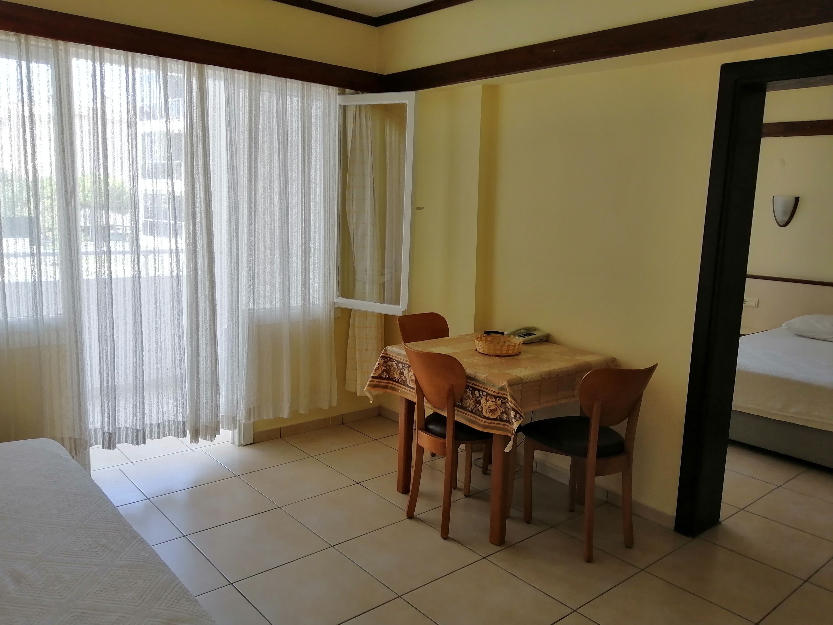 Habitación de hotel con cama, mesa de comedor con sillas, ventana con cortinas translúcidas y vistas al exterior.