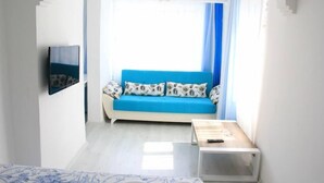 A room with a blue sofa, a TV mounted on the wall, a small table, and a window with curtains. - Mavi Butik Apart (Cesme)