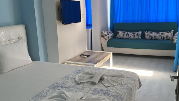 A hotel room with a bed, a sofa, a TV, and a window with blue curtains. - Mavi Butik Apart (Cesme)