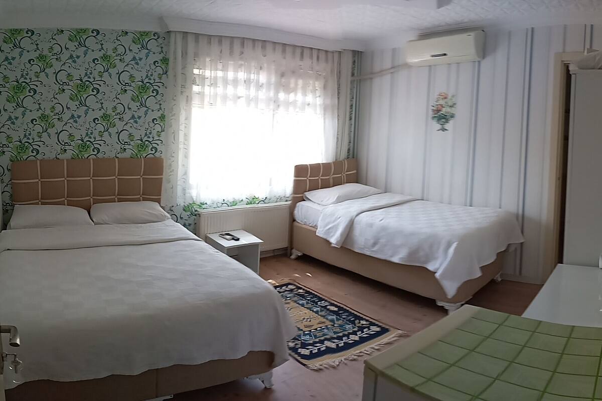 Habitación de hotel con dos camas, una mesita de noche, un radiador, una ventana con cortinas, aire acondicionado instalado en la pared y un baño con ducha y lavabo.