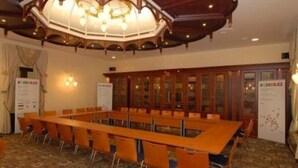 Een vergaderruimte met een lange tafel, stoelen, houten kasten en een decoratief plafond.
