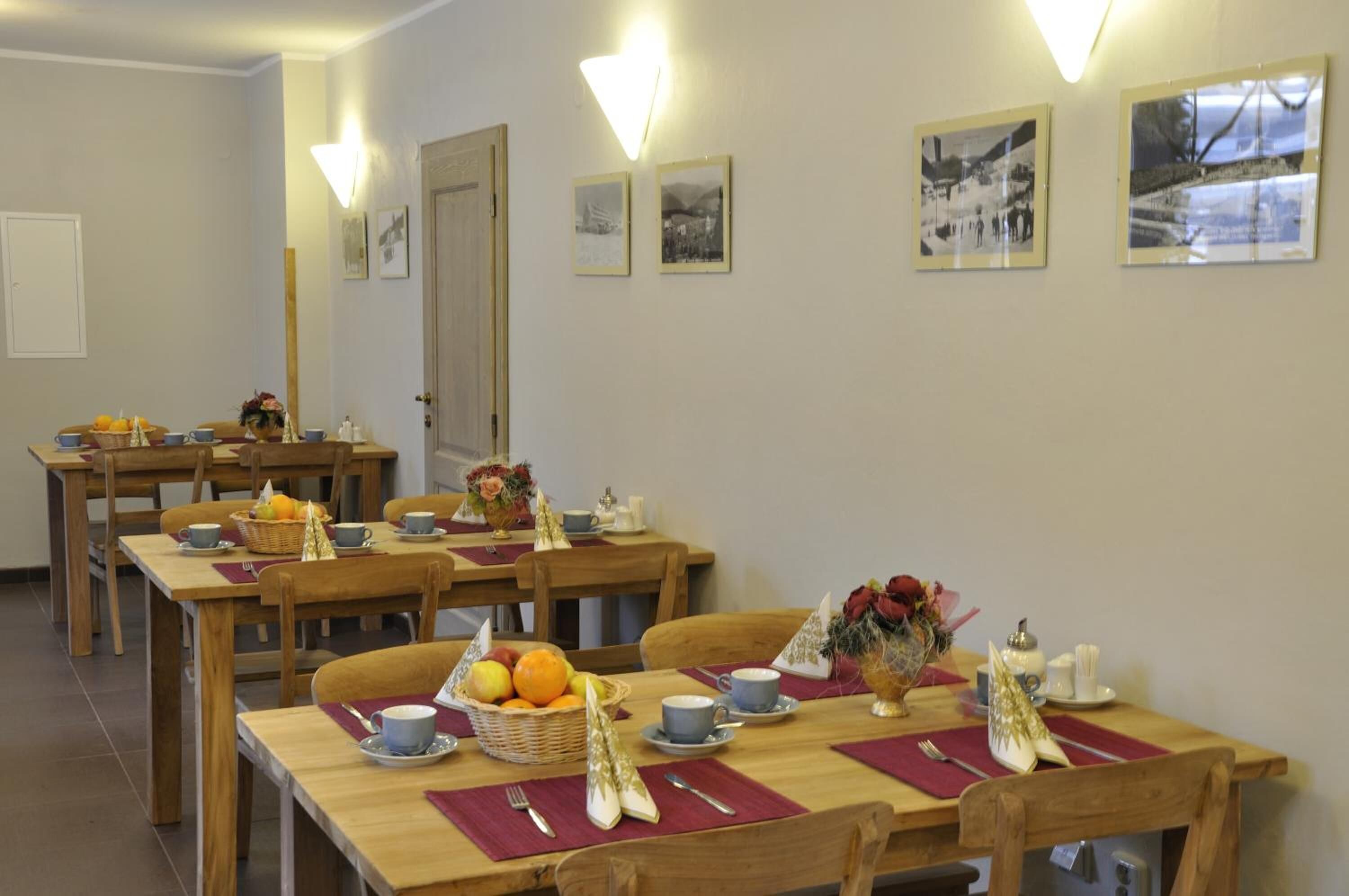 A dining area with wooden tables and chairs, each set with a placemat, a basket of fruit, and a vase of flowers.