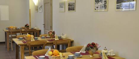 A dining area with wooden tables and chairs, each set with a placemat, a basket of fruit, and a vase of flowers.