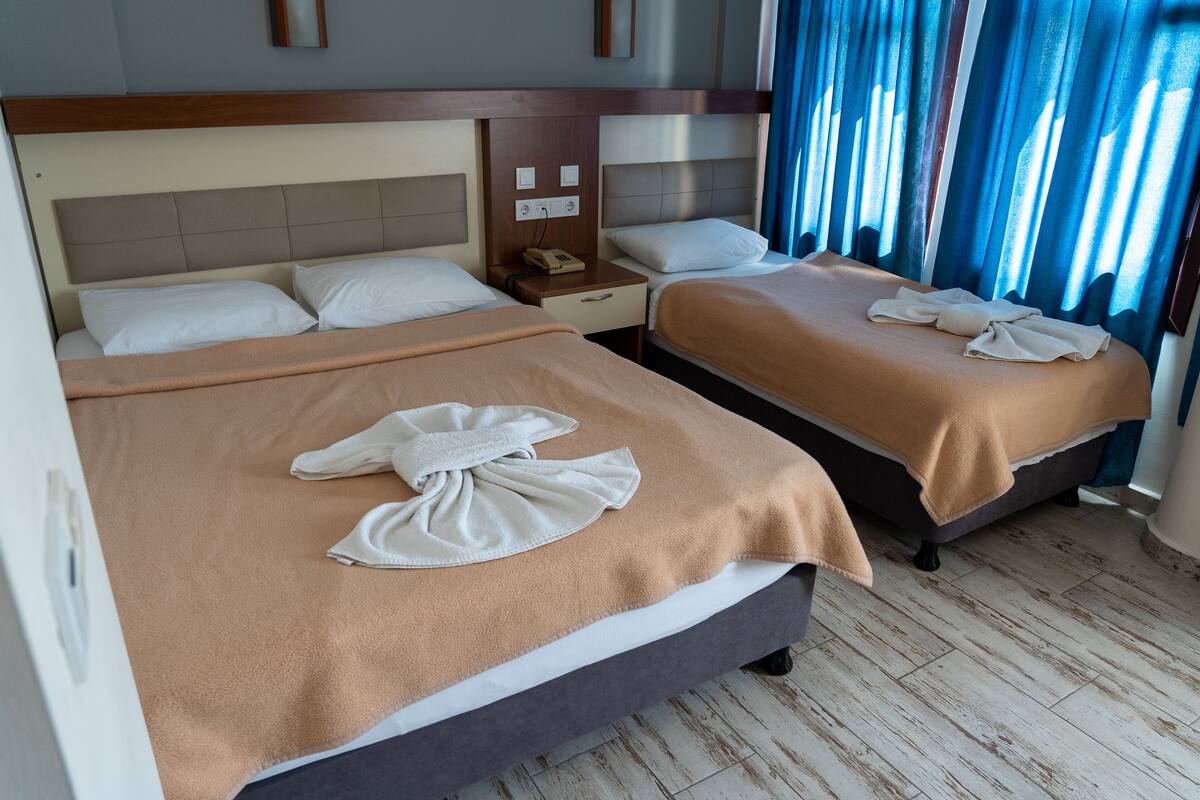 A hotel room with two beds, each with a brown blanket and a towel. There is a nightstand with a phone and a wall socket.