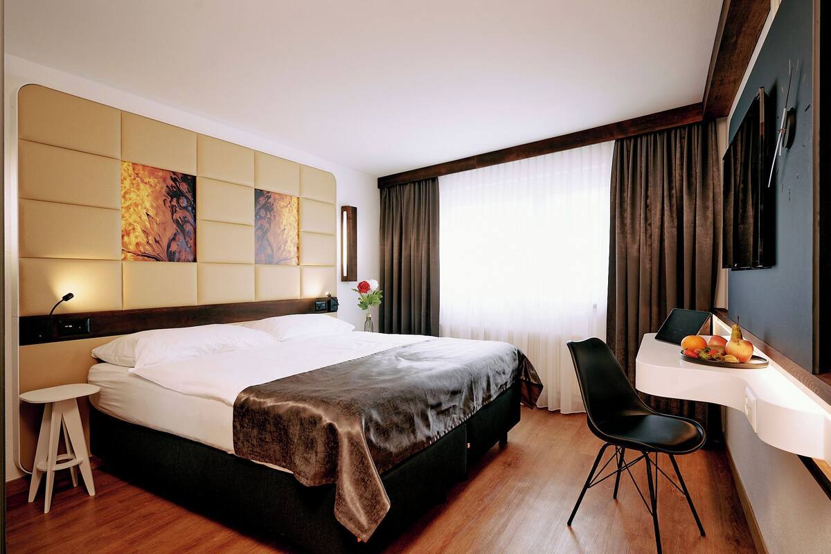 Ein modernes Hotelzimmer mit einem großen Bett, einem Schreibtisch mit Stuhl und Blick ins Freie durch große Fenster.