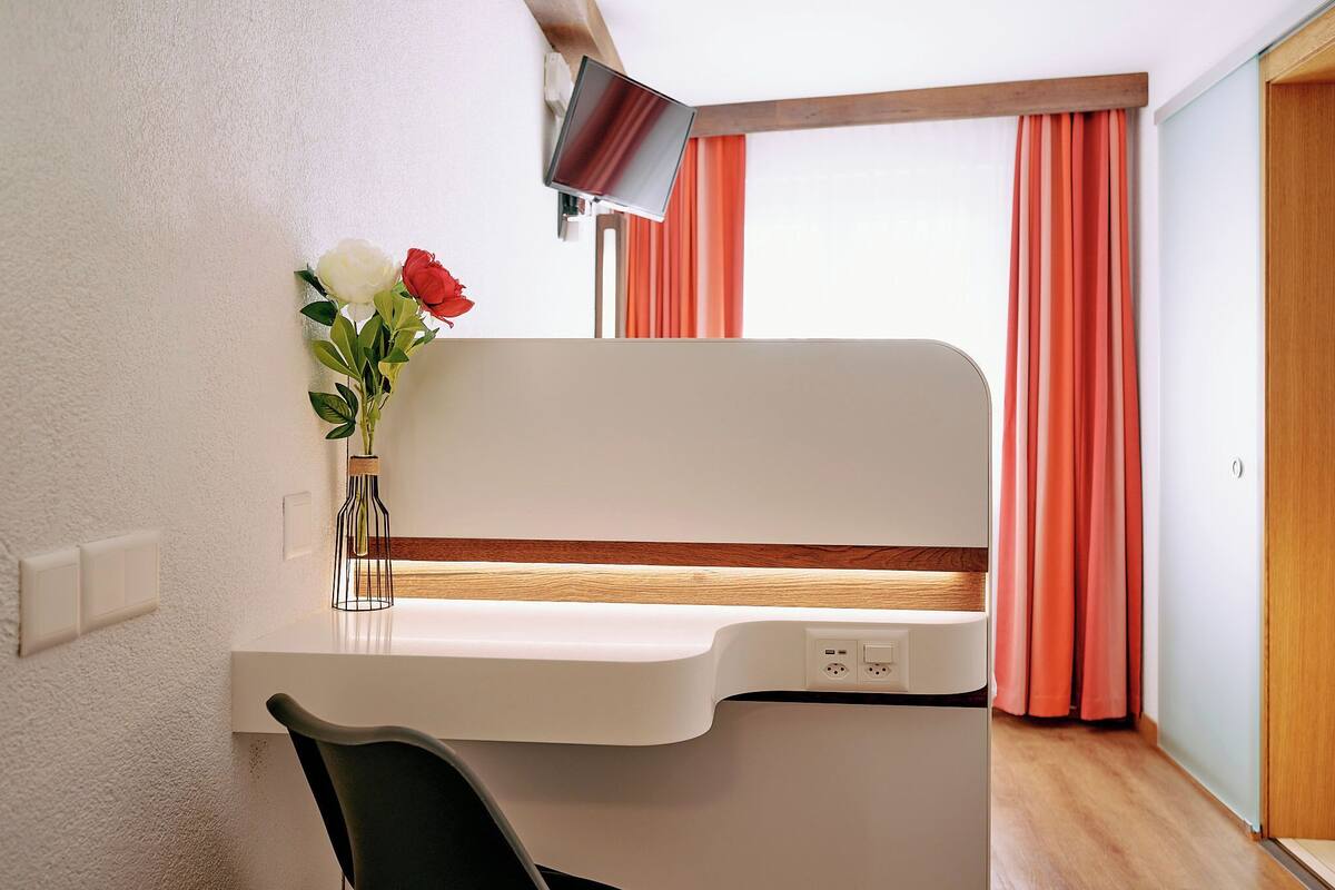 Ein gemütlicher Raum mit weißem Schreibtisch, schwarzem Stuhl, kleiner Blumenvase, Wandfernseher und Fenster mit orangefarbenen Vorhängen.