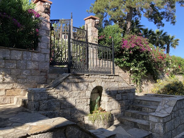 A stone staircase leading to a gated entrance with a wrought iron gate, surrounded by lush greenery and flowering plants.