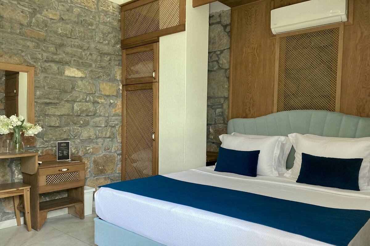 A bedroom with a stone wall, a wooden nightstand with a vase of flowers, a bed with white and blue bedding, and a wooden headboard.