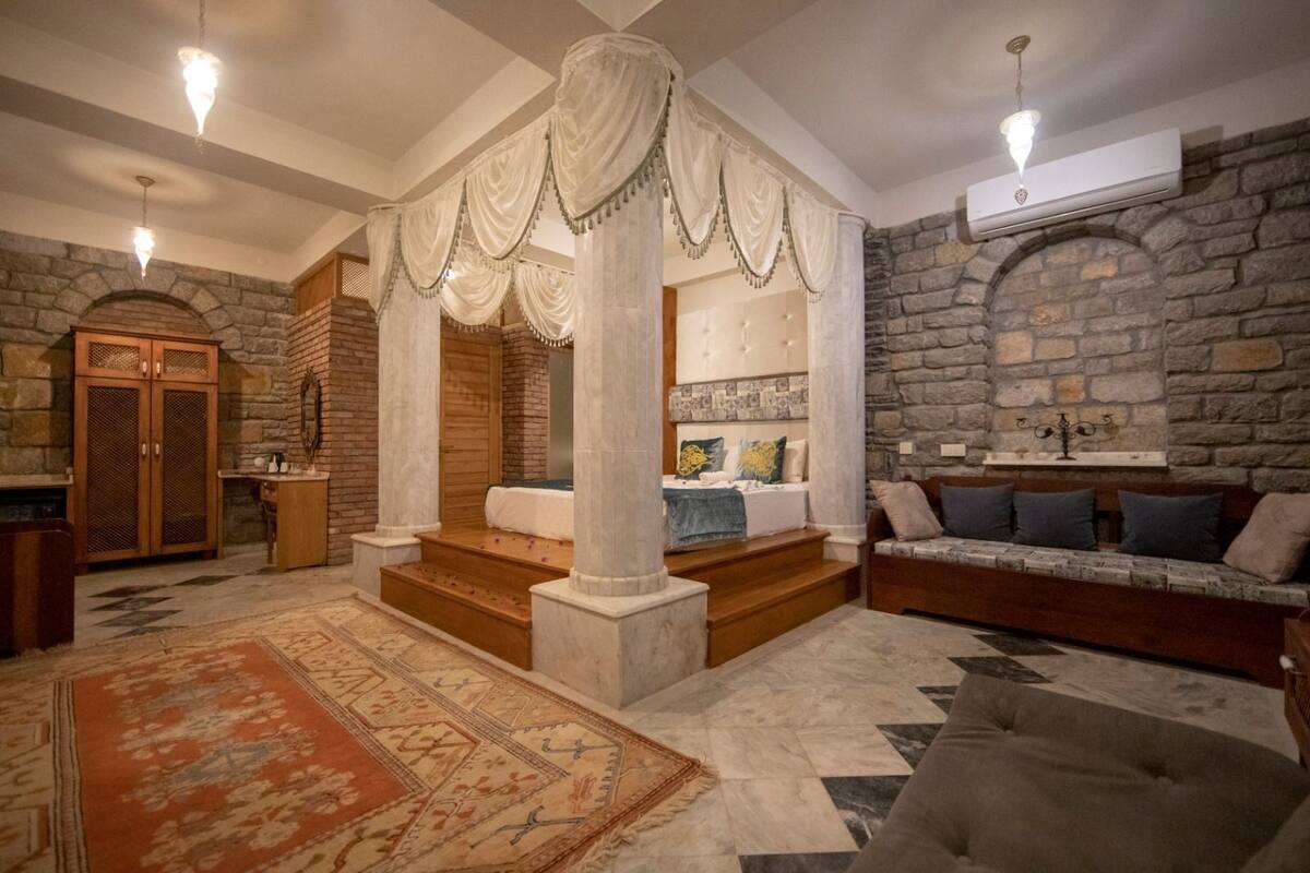A spacious living room with a stone wall, a wooden sofa, a patterned rug, and a staircase leading to a bedroom.