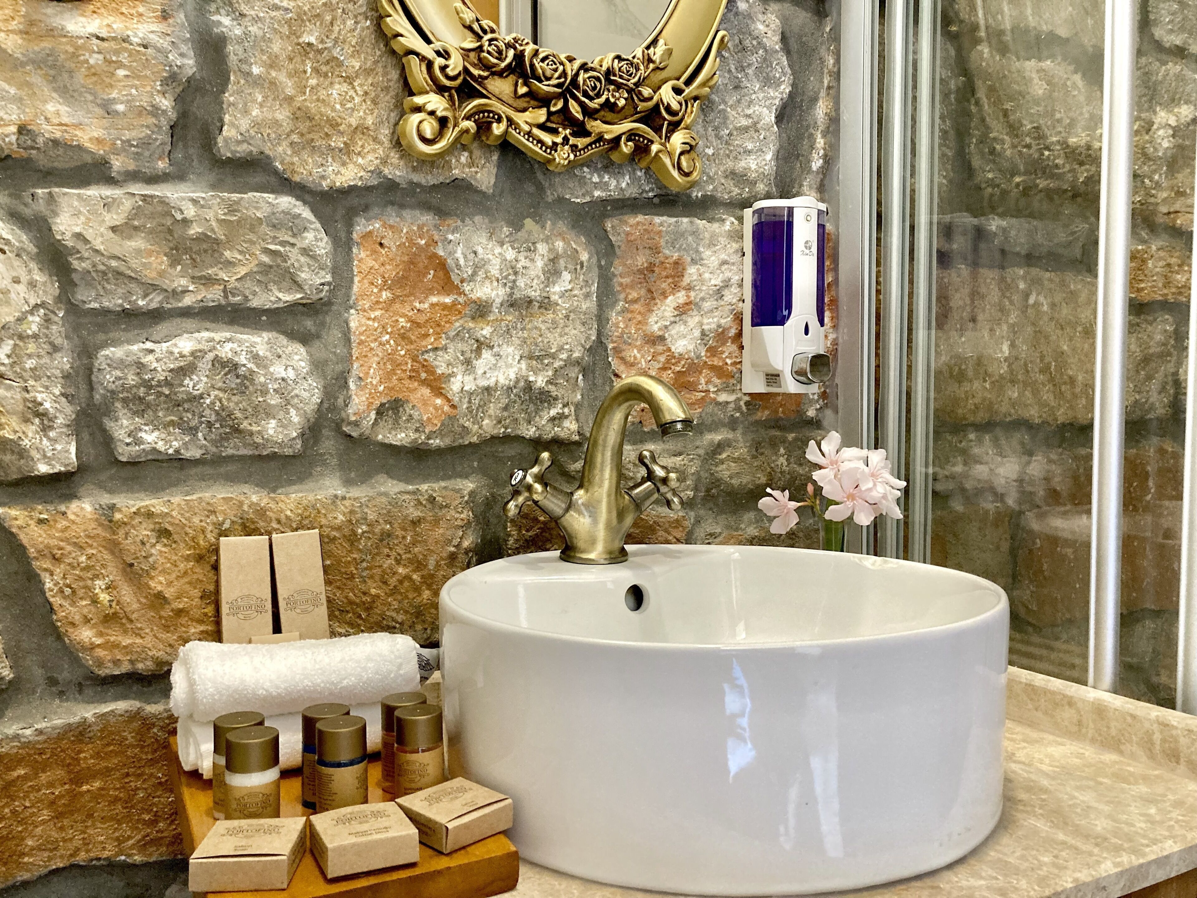 Ein Badezimmer mit Steinwand, goldenem Wasserhahn, weißem Waschbecken und Pflegeprodukten auf einer Ablagefläche.