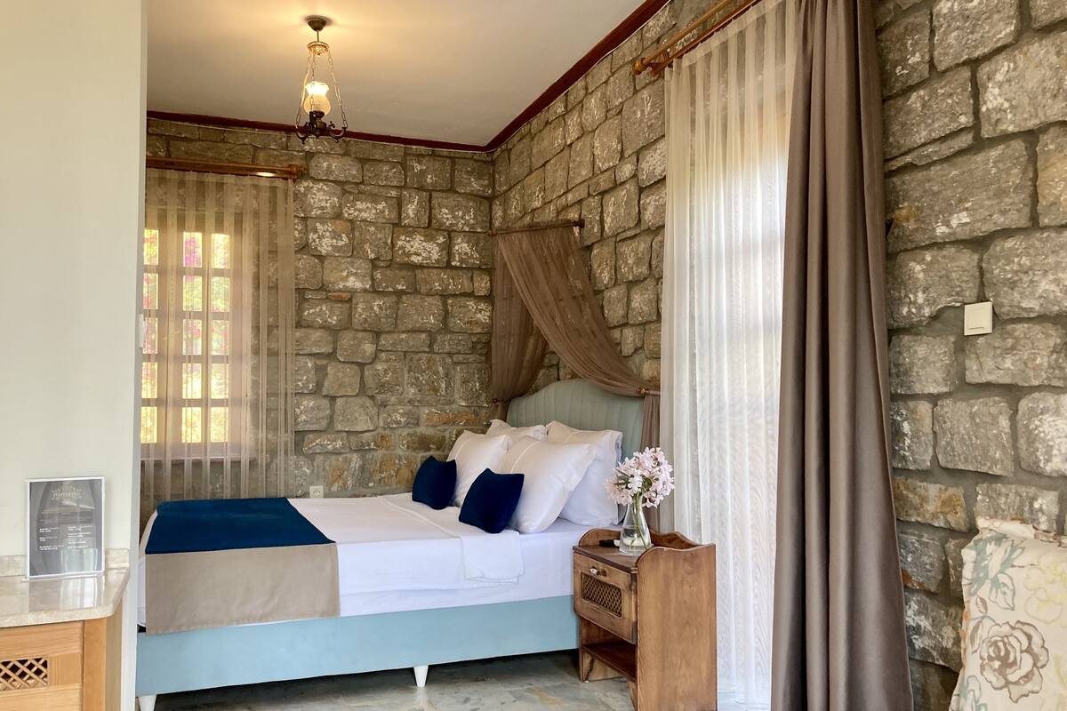 A bedroom with a stone wall, a bed with white bedding and blue pillows, a wooden nightstand with a vase, and a hanging light fixture.