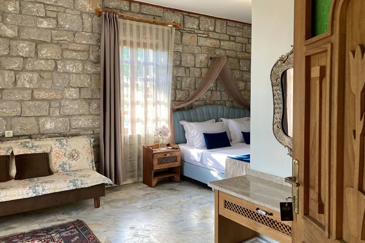 A bedroom with a stone wall, a bed with a canopy, a wooden nightstand, a sofa, and a small table.