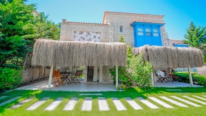 Un bâtiment en pierre avec une porte bleue, deux constructions à toits de chaume et un espace pavé avec des chaises et des tables.