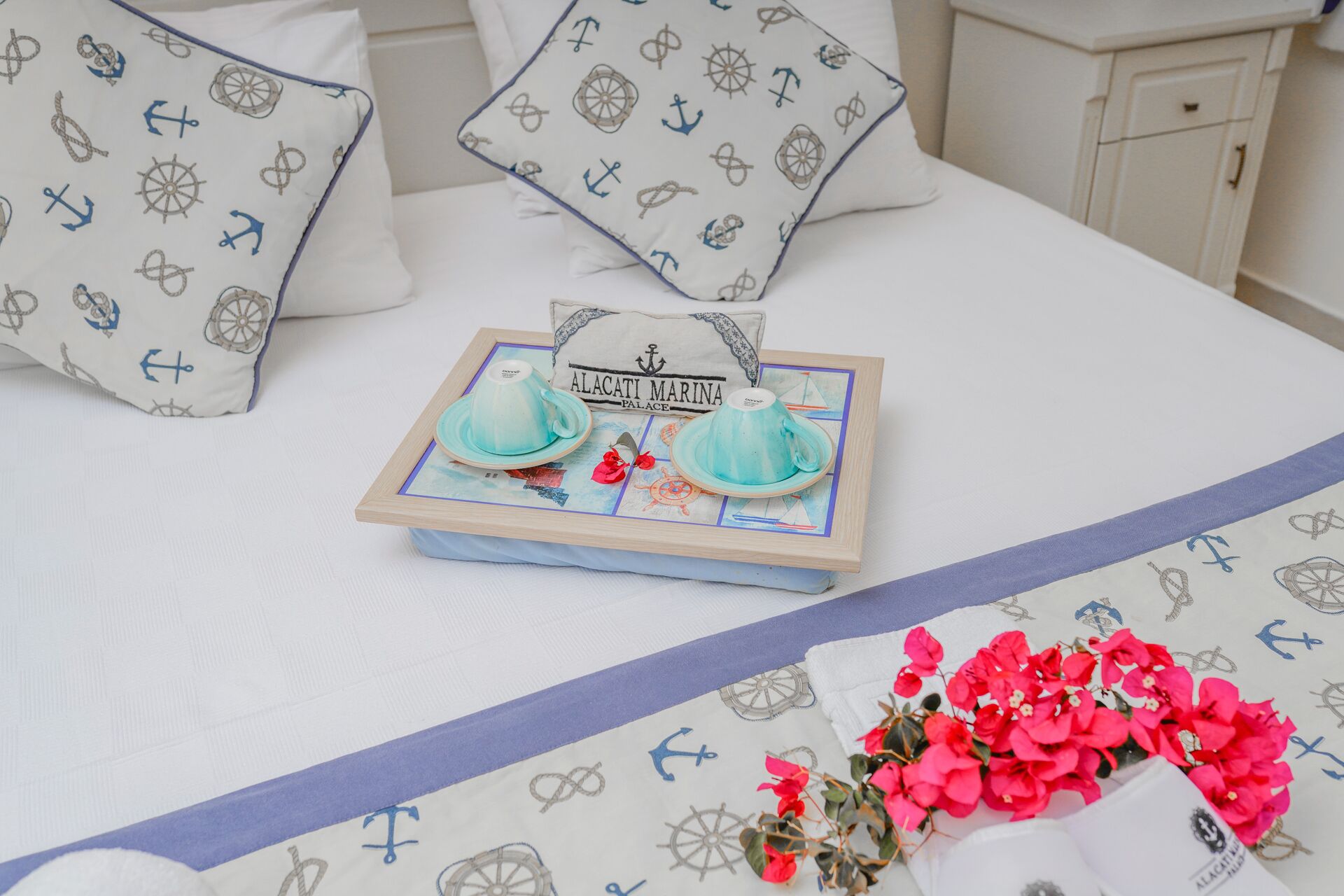 Una cama bien hecha con almohadas a juego, una bandeja con tazas de té y una almohada decorativa con el texto 'ALACATI MARINA PALACE'.