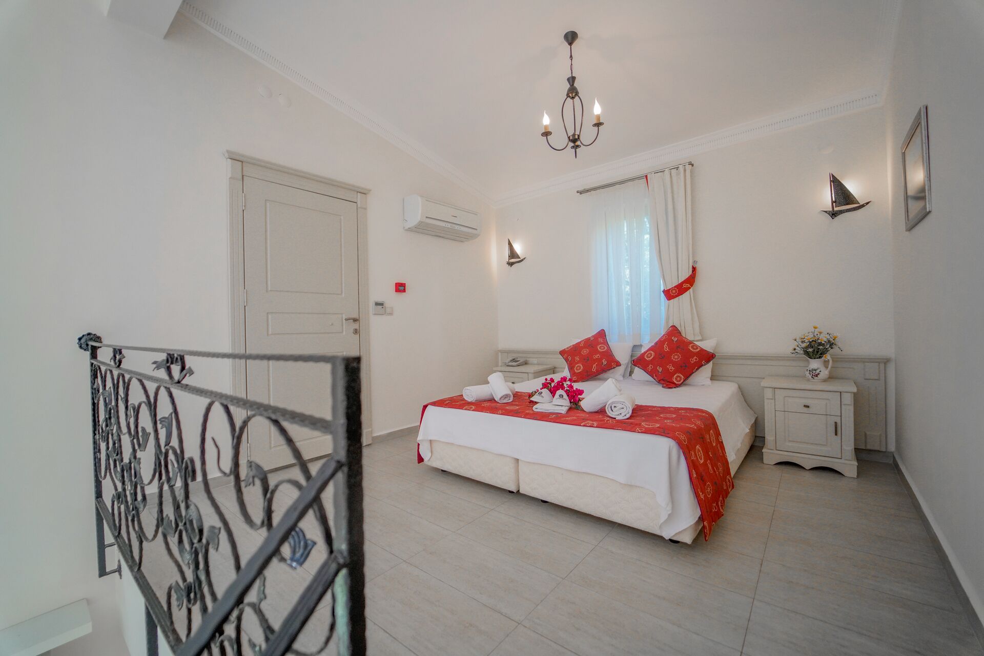 A hotel room with a bed, a nightstand, a lamp, a chandelier, and a window with curtains.