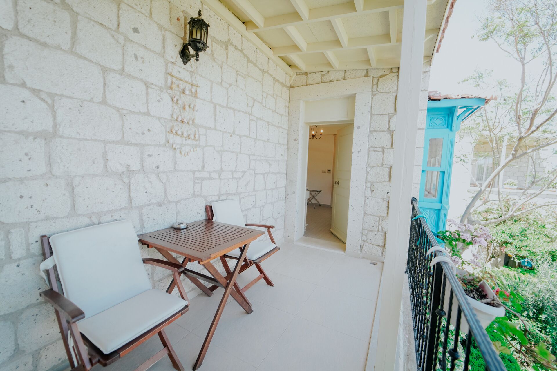 A balcony with white brick walls, a wooden table and chairs, a hanging lantern, and a view of a garden with a blue structure.