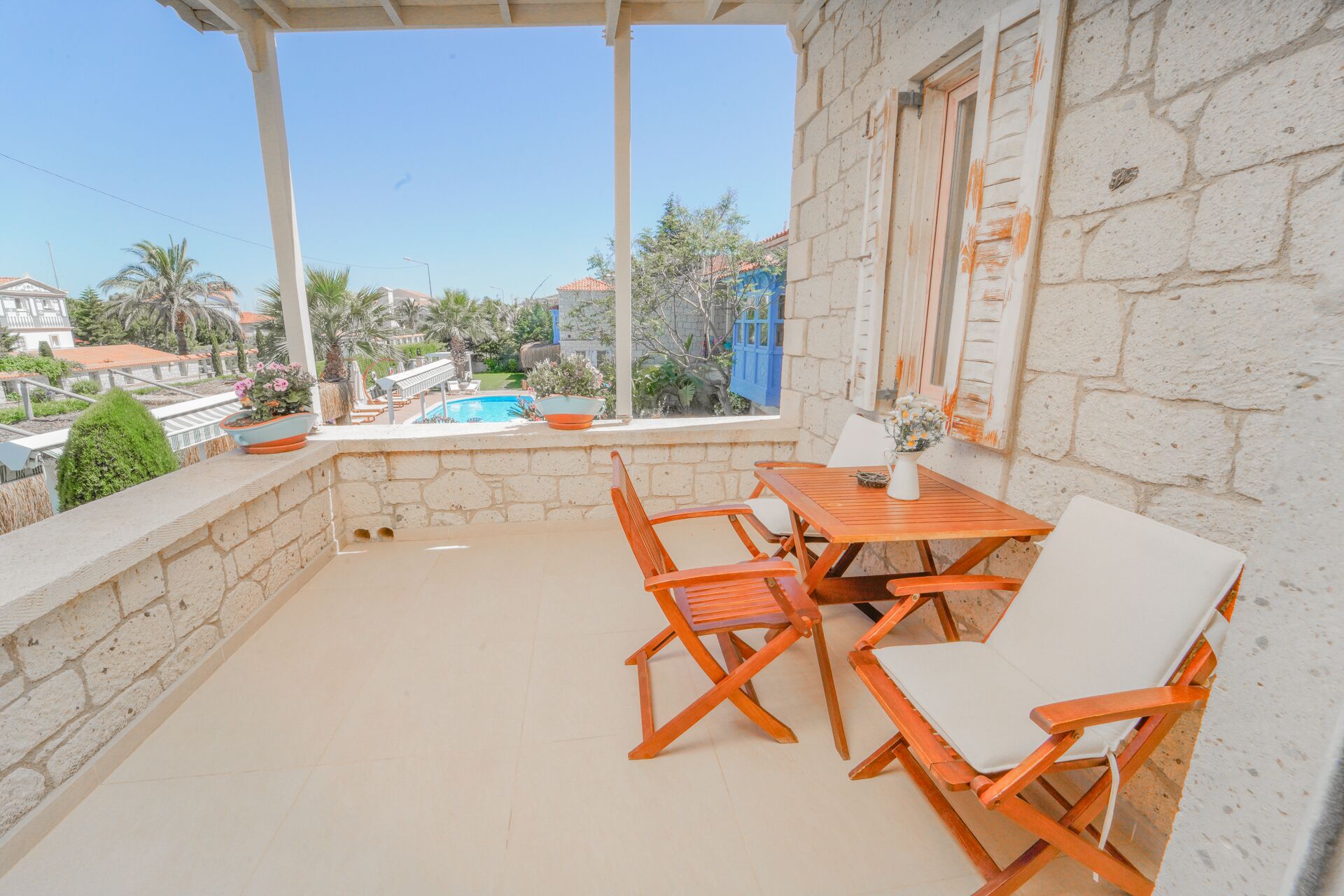 Un balcon avec une table et des chaises en bois, une vue sur une piscine et, au loin, un bâtiment en pierre.