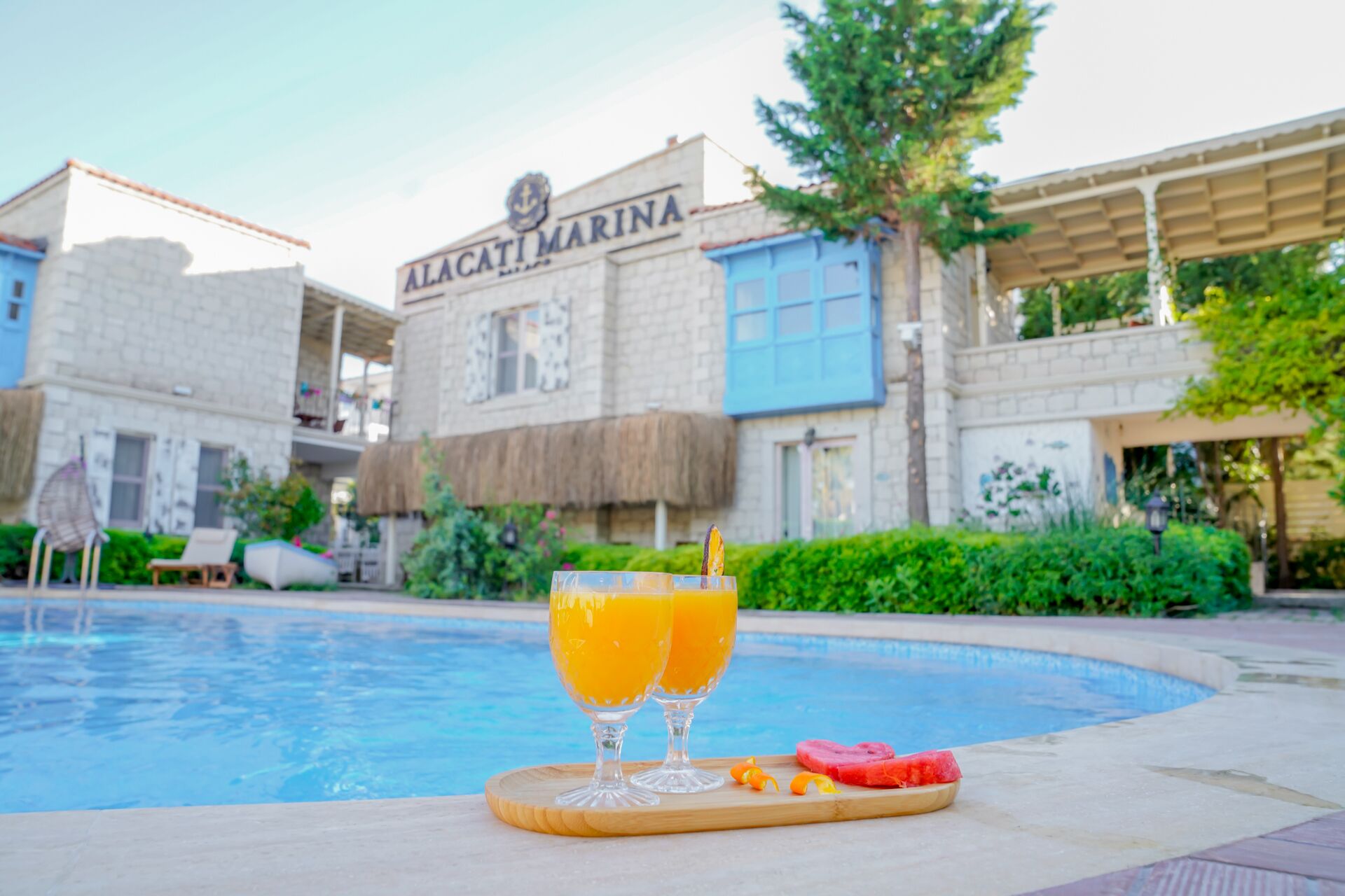 Une zone de piscine avec deux verres de jus d’orange et des tranches de pastèque disposées sur une planche en bois. En arrière-plan, on aperçoit un bâtiment portant l’inscription « ALACATI MARINA ».