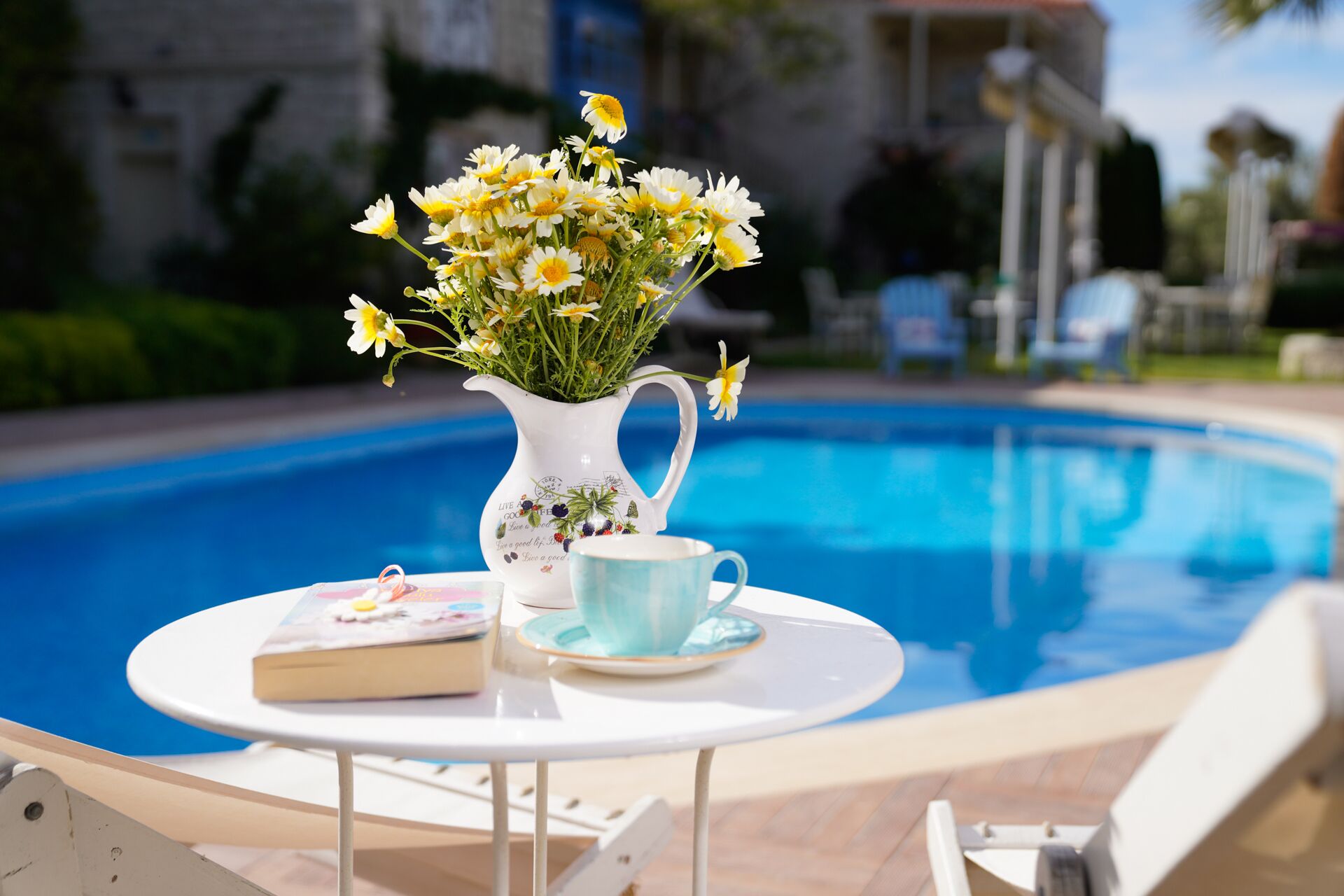 Escena junto a la piscina con una jarra blanca de flores, una taza azul y un libro sobre una mesita.