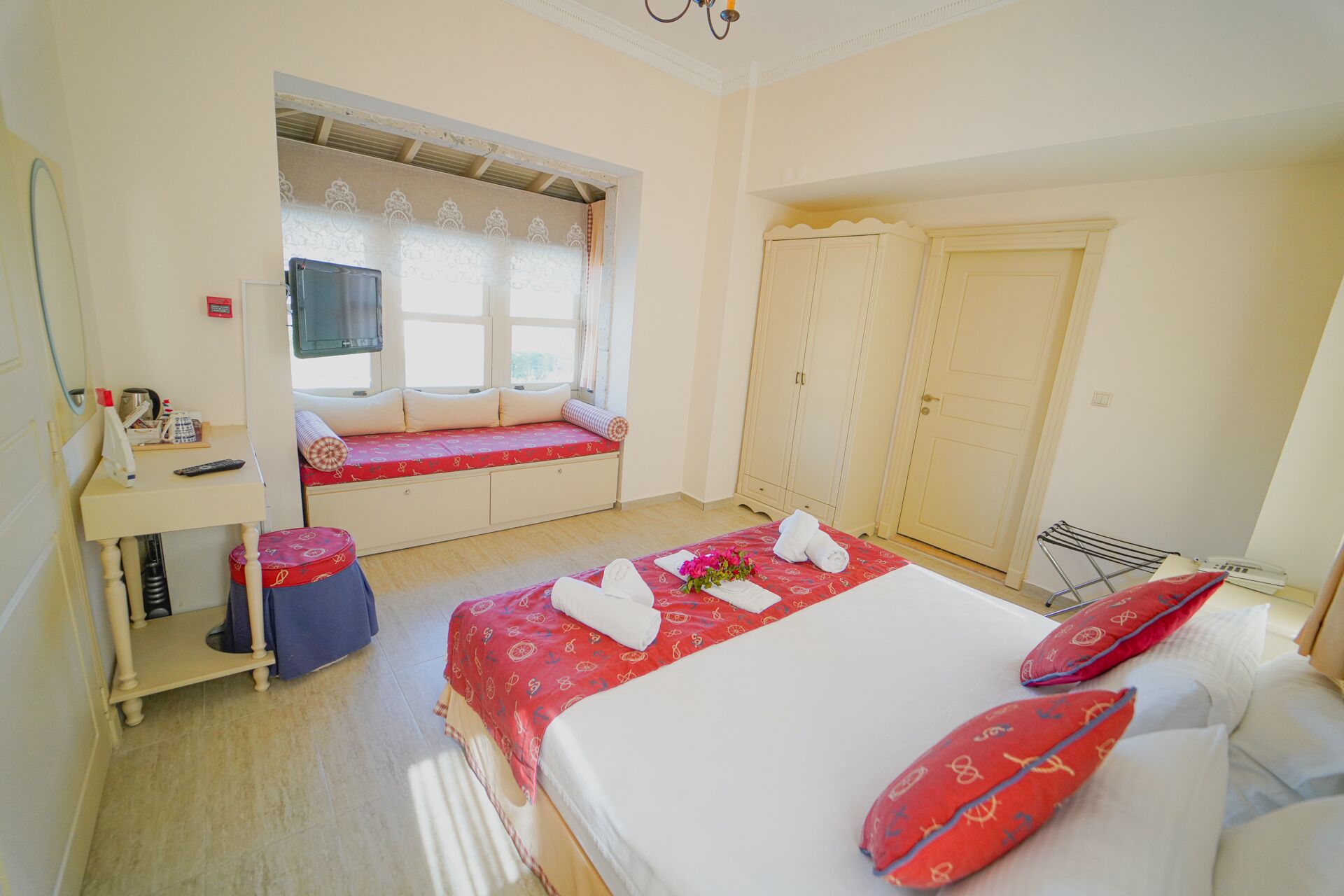 A hotel room with a bed, a window seat, a wardrobe, and a desk with a chair.