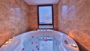Una bañera grande y ovalada con pétalos de rosa flotando en el agua, rodeada de velas, y una ventana que ofrece vistas al cielo.