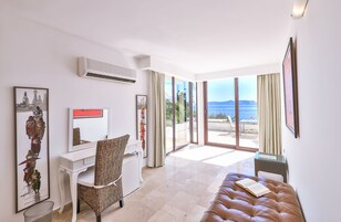 A modern hotel room with a desk, chair, and a large bed. There are multiple framed pictures on the walls and a view of the sea through large windows.