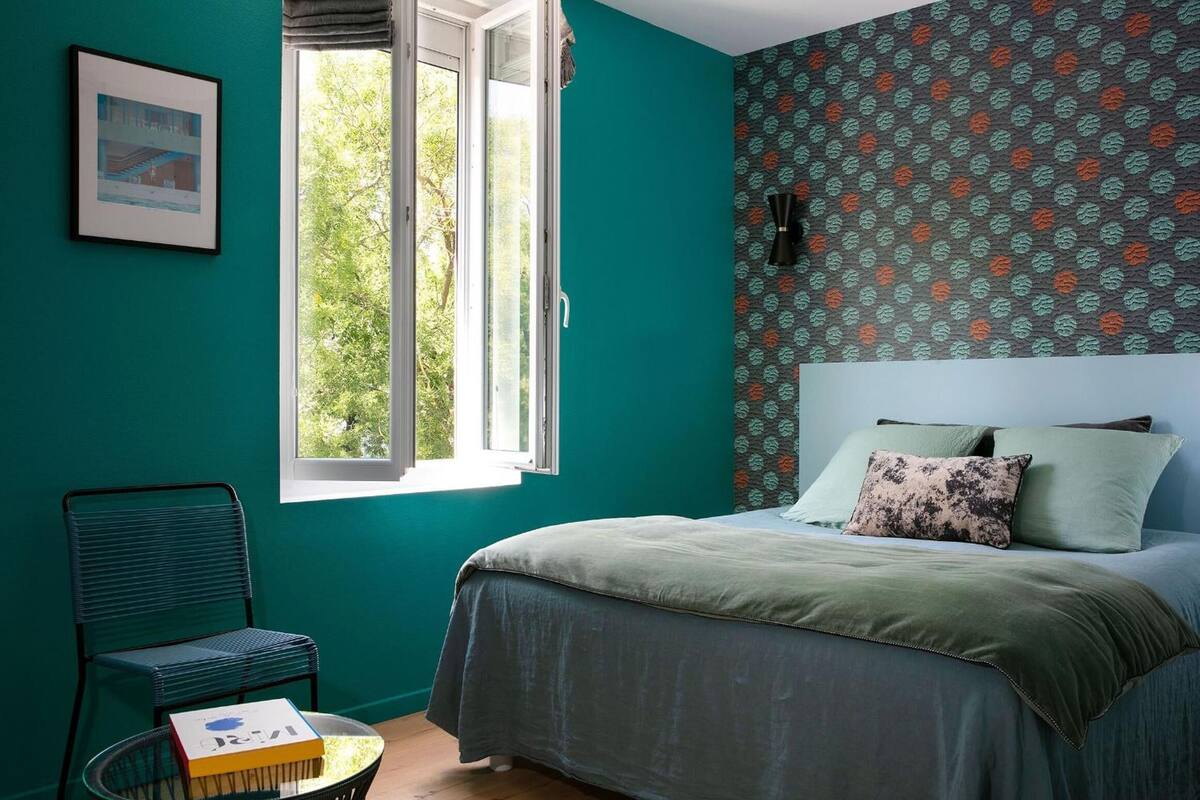 Un dormitorio con paredes azul verdosas, una cama con ropa de cama verde, una ventana con vista a árboles y un papel tapiz estampado.