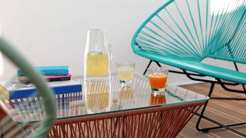 Ein Glastisch mit zwei Krügen Getränke und einem Glas Orangensaft, umgeben von bunten Büchern und einem Türkis-farbenen Stuhl.